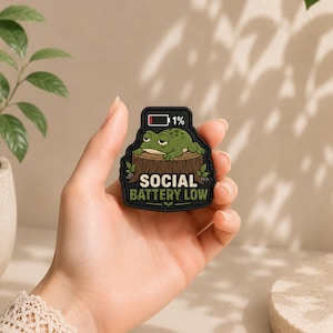 Puede incluir: Un pin de goma negro con una rana verde de dibujos animados descansando sobre un tocón de árbol marrón. Sobre la rana hay un icono de batería al 1%. El texto SOCIAL BATTERY LOW está impreso en letras blancas y verdes en la parte inferior.