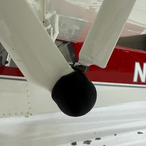 May include: A black, rounded protective cap fitted over the lower joint of two white metal landing gear struts on an aircraft. The aircraft fuselage in the background is white with a red horizontal stripe and a partial letter N visible in white.