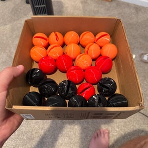 Pode incluir: Uma caixa de papelão contendo uma coleção de objetos esféricos nas cores laranja, vermelho e preto. Cada esfera apresenta uma única fenda vertical cortada na superfície superior. Os objetos estão dispostos em fileiras dentro do recipiente.