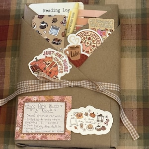 May include: A brown paper-wrapped book package tied with a gingham ribbon, decorated with autumn-themed stickers, a yellow pen labeled YOU'VE GOT THIS, and a note titled Blind Date With A Book describing a fall romance set in Connecticut.