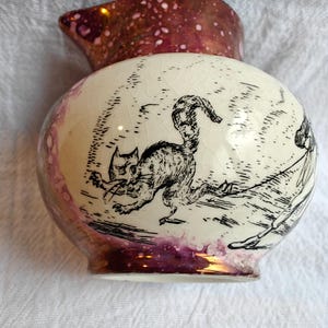 May include: A ceramic creamer pitcher with a cream-colored base featuring a fine crackle glaze finish. The body is decorated with a black ink illustration of a cat with an arched back. The rim and base have a metallic, iridescent purple luster.