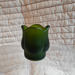 Peut inclure: Un vase en verre vert foncé avec une finition mate et givrée. Le vase présente une forme distincte rappelant une tulipe avec des bords incurvés en forme de pétales formant le rebord. La base est cylindrique et solide, s'élargissant vers le corps.