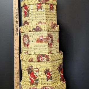Könnte beinhalten: Ein Stapel aus fünf sechseckigen Geschenkschachteln aus Pappe mit gelbem Hintergrund, Notenblättern und Weihnachtsmotiven wie dem Weihnachtsmann, Kränzen und Zügen. Ein Holzlineal steht daneben und zeigt eine Höhe von bis zu 79 cm an.