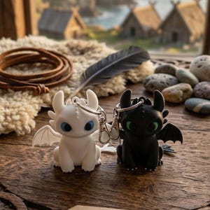 May include: Two dragon keychains, one white with blue eyes and one black with green eyes, each featuring small wings and a metal clasp. A wooden tag is present with a compass icon and the text DRAGON BUDDY KEYCHAINS printed in black ink.