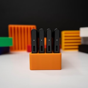 May include: Four slim black rectangular electronic devices with a single small white dot and a port on the bottom edge, standing upright in a bright orange rectangular plastic holder with four slots, arranged in a neat row on a white surface.
