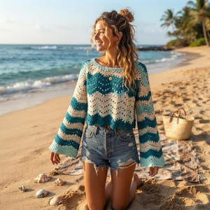 Puede incluir: Un suéter de ganchillo de manga larga con un patrón de ondas en rayas horizontales alternas de color verde azulado, azul claro y crema. El suéter tiene un cuello redondo, mangas acampanadas sueltas y un dobladillo corto con pantalones cortos de mezclilla.