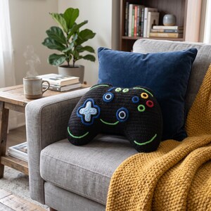 May include: A black crochet pillow shaped like a video game controller. The pillow features a light blue directional pad, blue and green circular buttons, and a green stitched smile. The texture is consistent with yarn-based needlework.