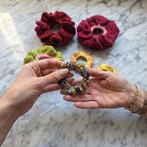May include: A collection of five handmade crochet hair scrunchies in various colors and textures. Two large scrunchies are deep red and magenta, while three smaller ones feature variegated green, yellow, and multi-colored yarn patterns.