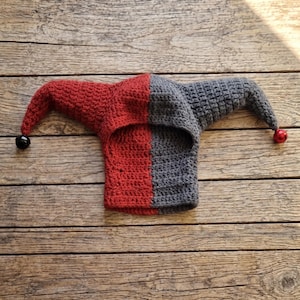 Puede incluir: Un gorro de bufón bicolor hecho de hilo de ganchillo, con un diseño dividido en rojo oscuro y gris oscuro. El gorro tiene dos solapas laterales largas y puntiagudas, cada una con un pequeño cascabel de metal redondo en el extremo.