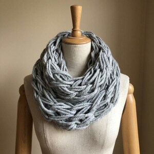 Peut inclure: Une écharpe infinie gris clair présentant un design tressé à mailles larges réalisé avec du fil épais. L'écharpe est drapée en plusieurs couches circulaires autour d'un buste de mannequin aux tons neutres, révélant un aspect tissé texturé.
