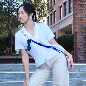 May include: A white short-sleeved collared shirt paired with beige trousers. A blue rope accessory with knots is worn across the torso, featuring a silver chain section and black sunglasses attached to the cord. The shirt has a button-down front.