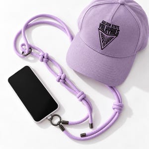 May include: A light purple baseball cap with the black text Golden State Valkyries above a triangular graphic, connected to a matching purple rope phone lanyard with dark metal rings and end caps, holding a black smartphone.