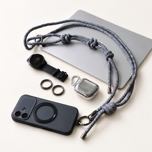 May include: A grey speckled rope lanyard with metal hardware, a black smartphone case with a circular magnetic ring, a black smartwatch, two small dark metal rings, a silver-toned wireless earbud charging case, and a grey tablet device.