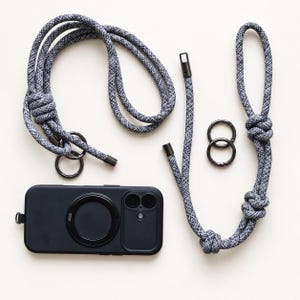 May include: A black smartphone case with a circular magnetic attachment, accompanied by two grey and white speckled rope lanyards featuring dark metal hardware and two separate dark metal rings for versatile attachment and carrying options.
