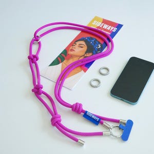 May include: A bright magenta rope phone lanyard featuring multiple decorative knots, silver-toned metal hardware, and a blue rectangular tag with the text SIDEWAYS. The lanyard includes a silver ring attachment and two additional loose silver rings.