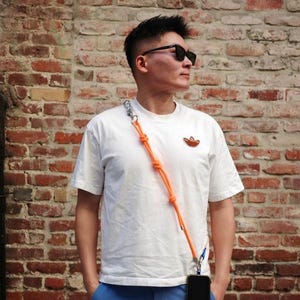 May include: A white short-sleeved t-shirt with an orange logo on the chest, paired with blue shorts. A bright orange rope phone lanyard with silver hardware is worn across the body, holding a black smartphone at the hip.