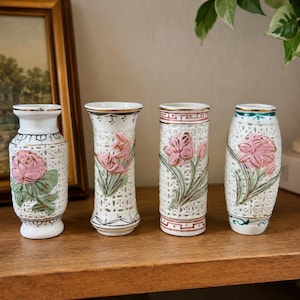 Peut inclure: Quatre petits vases en céramique blanche présentant des motifs complexes en treillis ajouré et des reliefs de fleurs roses avec des feuilles vertes. Chaque vase est rehaussé de bordures dorées sur les cols et les bases, avec des formes variées.