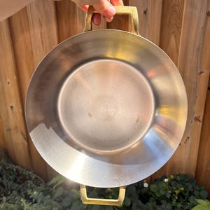 May include: A round, shallow metal pan with a brushed silver finish and two gold-toned, rectangular metal handles attached to opposite sides. The pan features a smooth, circular interior surface and a wide, flat rim, presenting a clean, metallic design.