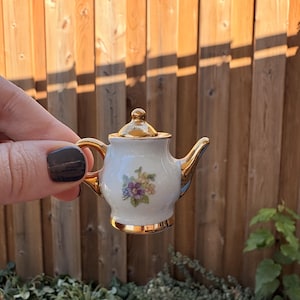 May include: A miniature white porcelain teapot featuring a delicate floral pattern with purple and yellow flowers. The teapot has a shiny gold-coloured handle, spout, base, and lid, creating a reflective metallic finish on these specific parts.