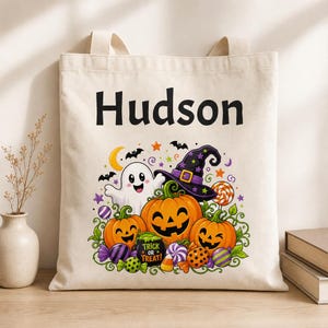 Peut inclure: Un sac fourre-tout en toile beige portant le nom Hudson en noir au-dessus d'une illustration colorée d'Halloween représentant un fantôme blanc, trois citrouilles, un chapeau de sorcière, des bonbons et des chauves-souris.