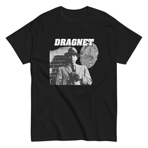 May include: A black short-sleeved t-shirt featuring a white graphic print of a detective in a hat and suit standing before a brick wall. The text DRAGNET appears above the figure, alongside an illustration of a Los Angeles Police badge number 714.