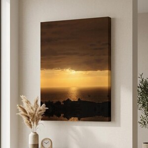 Puede incluir: Un lienzo rectangular para pared que muestra una puesta de sol sobre el océano con nubes oscuras arriba y la silueta de una ciudad costera abajo. La escena presenta una luz dorada que se refleja en el agua, creando un alto contraste visual.