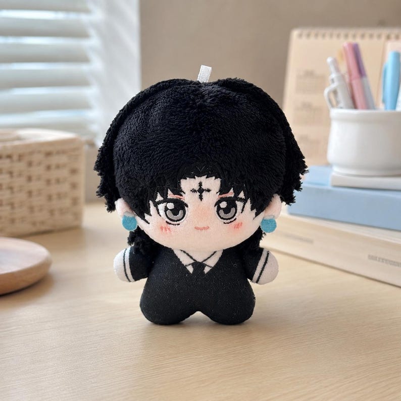 Pu&ograve; includere: Una bambola di peluche con capelli neri corti, grandi occhi grigi e un piccolo simbolo a croce sulla fronte. La bambola indossa un completo nero con dettagli bianchi sul colletto e piccoli orecchini circolari blu ai lati.