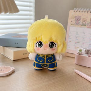 Pu&ograve; includere: Una bambola di peluche con capelli giallo brillante, grandi occhi verdi e un piccolo sorriso. La bambola indossa un completo blu con finiture gialle e ha un piccolo occhiello sulla parte superiore della testa. Il peluche &egrave; in tessuto morbido.