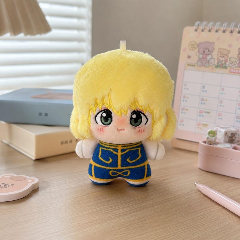 Pu&ograve; includere: Una bambola di peluche con capelli giallo brillante, grandi occhi verdi e un piccolo sorriso. La bambola indossa un completo blu con finiture gialle e ha un piccolo occhiello sulla parte superiore della testa. Il peluche &egrave; in tessuto morbido.