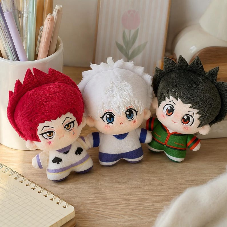 Pu&ograve; includere: Tre bambole di peluche con design ispirati agli anime. La bambola a sinistra ha i capelli rossi e un completo bianco con simboli di picche neri. Quella centrale ha i capelli bianchi e un completo blu e bianco. Quella a destra ha i capelli scuri e un completo verde.