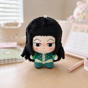 Pu&ograve; includere: Una bambola di peluche che raffigura un personaggio con folti capelli neri, pelle pallida e un'espressione neutra. La bambola indossa un completo verde con dettagli gialli e un colletto bianco. Ha un piccolo occhiello bianco sulla sommit&agrave; del capo.