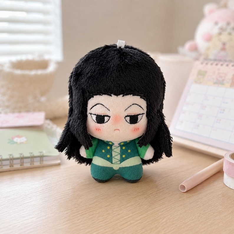 Pu&ograve; includere: Una bambola di peluche che raffigura un personaggio con folti capelli neri, pelle pallida e un'espressione neutra. La bambola indossa un completo verde con dettagli gialli e un colletto bianco. Ha un piccolo occhiello bianco sulla sommit&agrave; del capo.