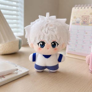 Pu&ograve; includere: Una bambola di peluche con capelli bianchi corti e a punta, grandi occhi blu e guance rosate. La bambola indossa una maglietta bianca con accenti blu sulle maniche e una parte inferiore blu, e include un piccolo occhiello bianco sulla testa.