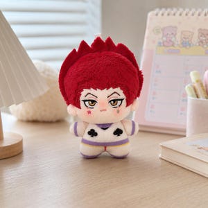 Pu&ograve; includere: Una bambola di peluche con capelli rosso vivo, occhi color ambra e un piccolo segno a forma di lacrima sotto l'occhio sinistro. La bambola indossa un completo bianco con simboli neri di fiori e picche sul petto, rifinito con bordi viola.