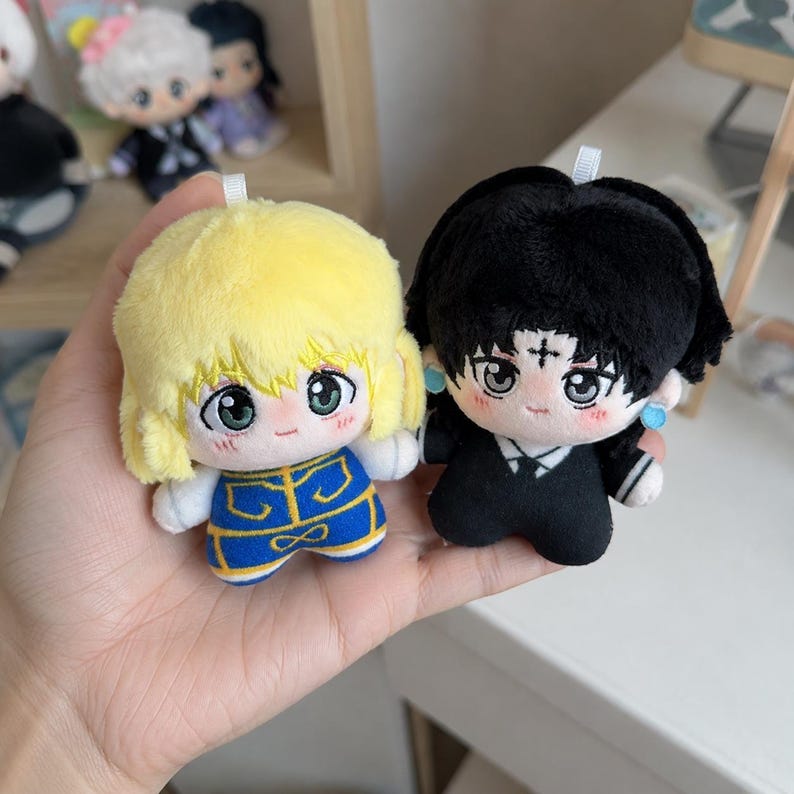 Pu&ograve; includere: Due piccole bambole di peluche con tratti del viso ricamati. Una bambola ha capelli giallo brillante e un vestito blu e oro. L'altra ha capelli neri, un simbolo a croce sulla fronte e un abito nero con orecchini circolari blu.