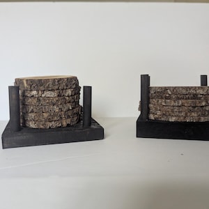 May include: Two sets of circular wood slice coasters stacked within black square wooden holders. Each holder features four vertical black posts at the corners to secure the natural-edged, light brown wood slices in a neat, vertical stack.