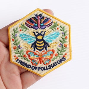 May include: A hexagonal embroidered patch with a cream background and a yellow border. The design features a central bee, a moth above, and a butterfly below, surrounded by green foliage and small flowers. The text reads FRIEND OF POLLINATORS.
