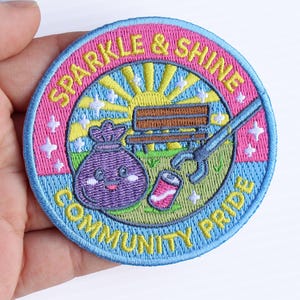 May include: A circular embroidered patch featuring a light blue border and a pink inner ring. The center depicts a smiling purple trash bag, a brown park bench, a yellow sun, and a blue grabber tool picking up a pink soda can. Text reads SPARKLE & SHINE COMMUNITY PRIDE.