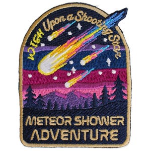 May include: An embroidered patch with a dark blue border featuring a night sky with pink and purple mountains, pine trees, and three shooting stars. The text reads "Wish Upon a Shooting Star" at the top and "METEOR SHOWER ADVENTURE" at the bottom.
