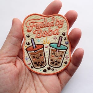 May include: An embroidered patch with a cream-colored background and an orange border. The text reads "Fueled by Boba" in orange cursive. Below the text are two cartoon boba tea cups with smiling faces, one with an orange lid and one with a teal lid.