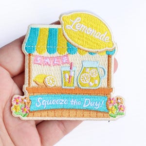 May include: An embroidered patch featuring a lemonade stand with a yellow lemon-shaped sign that reads Lemonade. The stand has a blue and yellow striped awning, a pink banner that says SALE, a glass of lemonade, and a pitcher. It says Squeeze the Day!