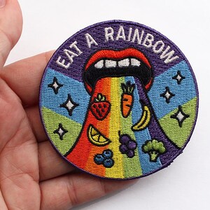 May include: A circular embroidered patch featuring a mouth with red lips and white teeth, emitting a rainbow stream containing a strawberry, carrot, banana, lemon slice, grapes, blueberries, and broccoli, with the text EAT A RAINBOW.