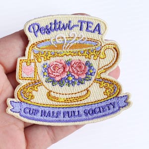 May include: An embroidered patch featuring a cream-colored teacup with pink roses, a yellow rim, and a tea bag with a pink heart. The text reads Positivi-TEA at the top and CUP HALF FULL SOCIETY on a light purple banner at the bottom.