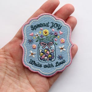 May include: A light blue embroidered patch with a scalloped pink border. The design features a glass jar filled with colorful geometric shapes, topped with flowers, and the text "Spread JOY" and "Write with Love" in dark blue thread.