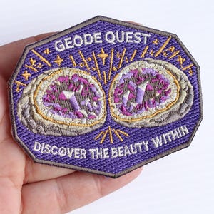 May include: An embroidered patch with a purple background featuring two split geode rocks with purple and white crystals. The text GEODE QUEST is at the top and DISCOVER THE BEAUTY WITHIN is at the bottom in white thread with gold star accents.