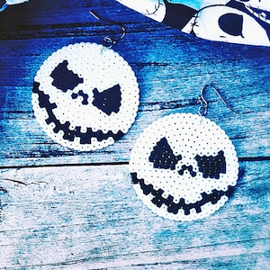 May include: Pair of round white earrings with a black Jack Skellington face design. The earrings are made of small beads and have silver-colored hooks. The background is a blue-toned wooden surface with a black and white patterned fabric.