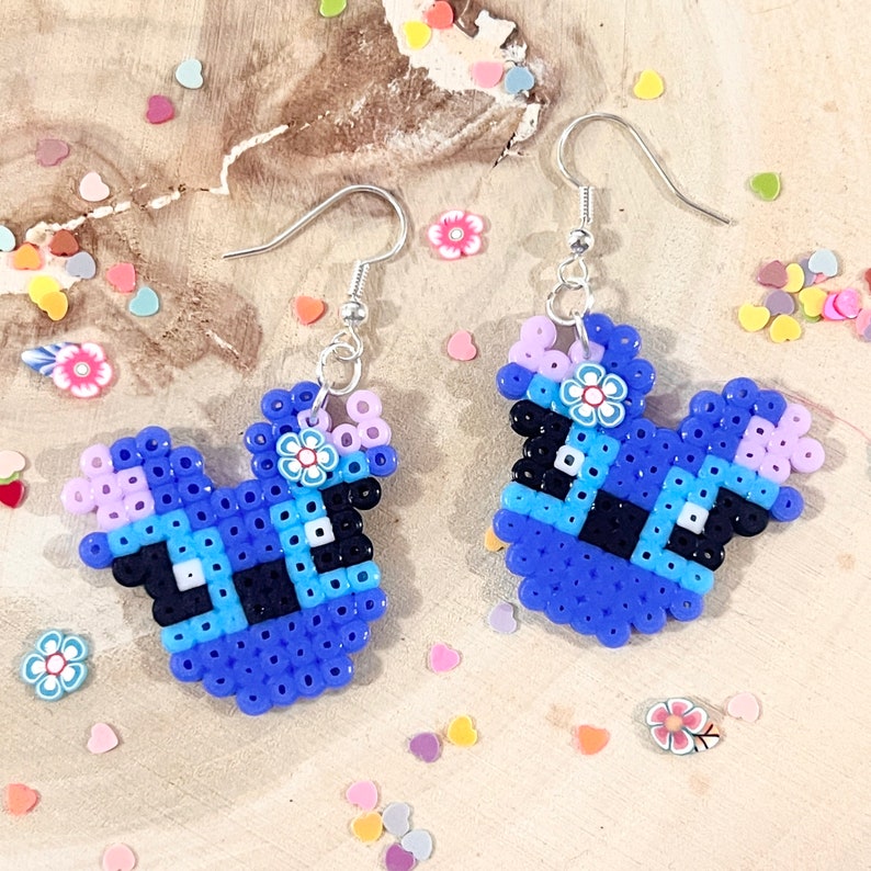 Puede incluir: Un par de pendientes de cuentas azules y moradas con forma de personaje de dibujos animados con una flor en la parte superior. Los pendientes est&aacute;n unidos a ganchos plateados.