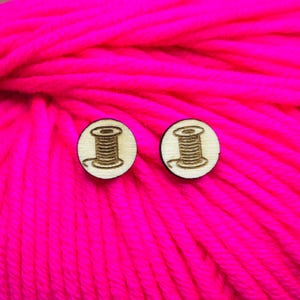 May include: A pair of wooden earrings with a spool of thread design. The earrings are a light brown color and are set against a bright pink background.