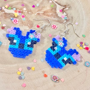 Puede incluir: Un par de pendientes de cuentas azules y moradas con un dise&ntilde;o de flores. Los pendientes est&aacute;n sobre una superficie de madera con confeti de colores.