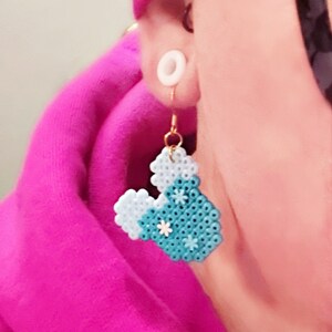 Puede incluir: Un par de pendientes en forma de coraz&oacute;n azul y blanco con flores blancas. Los pendientes est&aacute;n hechos de peque&ntilde;as cuentas y cuelgan de ganchos dorados.